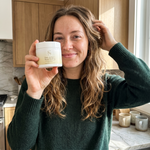 Young woman holding Pure Tallow+ Deep Healing jar in bright kitchen morning light