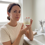 Woman holding Pure Tallow+ Deep Healing jar at bathroom vanity morning